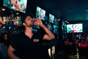 Le barman boit un shot devant les bouteilles et les écrans du bar